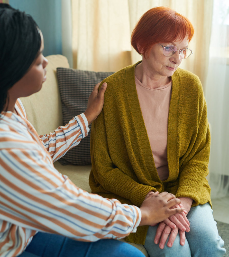 Depressed senior woman sitting on sofa with sad expression while caregiver talking to her and supporting her
