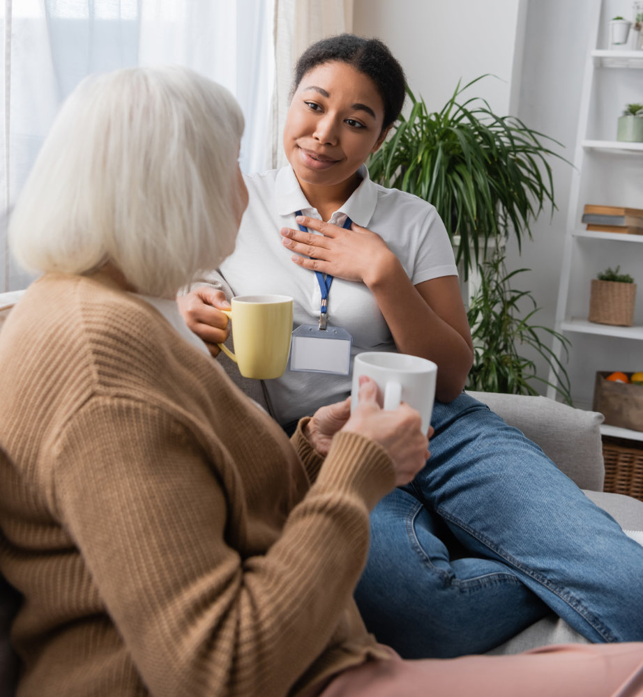 Compassionate multiracial care giver chatting with senior woman while having tea in living room.