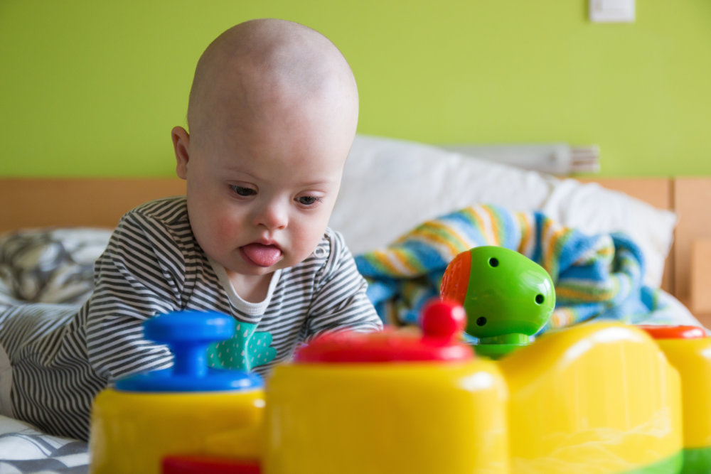 Cute baby boy with Down syndrome playing with toy on the bed in home bedroom