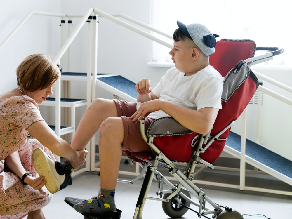 Mother helping disabled boy 17 years old in wheelchair with cerebral palsy to put on shoes, before going for a walk.