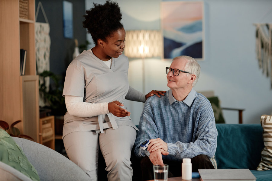 Black female caregiver talking to an elderly man while measuring oxygen saturation in blood with oximeter sitting on couch at home.
