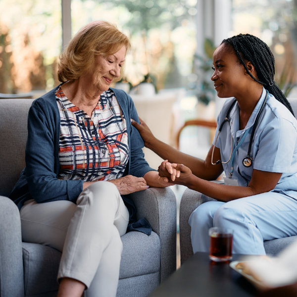 Sad senior woman holding hand with young African American nurse who is comforting her at home.