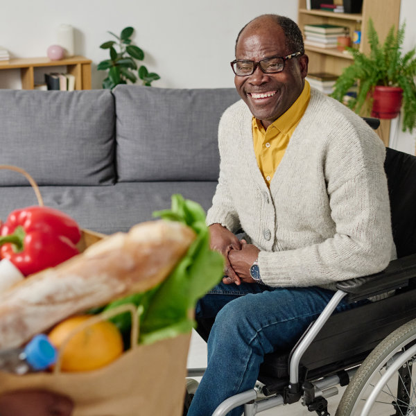 African happy man with disability sitting in wheelchair and meeting caregiver bringing groceries for him