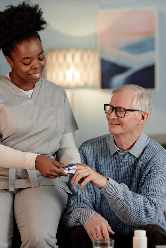 Supportive Black female social worker visiting upbeat elderly man checking up oxygen saturation in blood with pulse oximeter, while sitting together on couch providing medical assistance at home