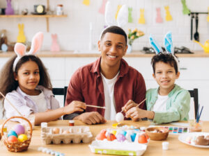 Happy family painting easter eggs wearing bunny ears. Smiling African American man, father and two kids sitting in the kitchen looking at camera.