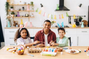 Happy family painting easter eggs wearing bunny ears. Smiling African American man, father and two kids sitting in the kitchen looking at camera.